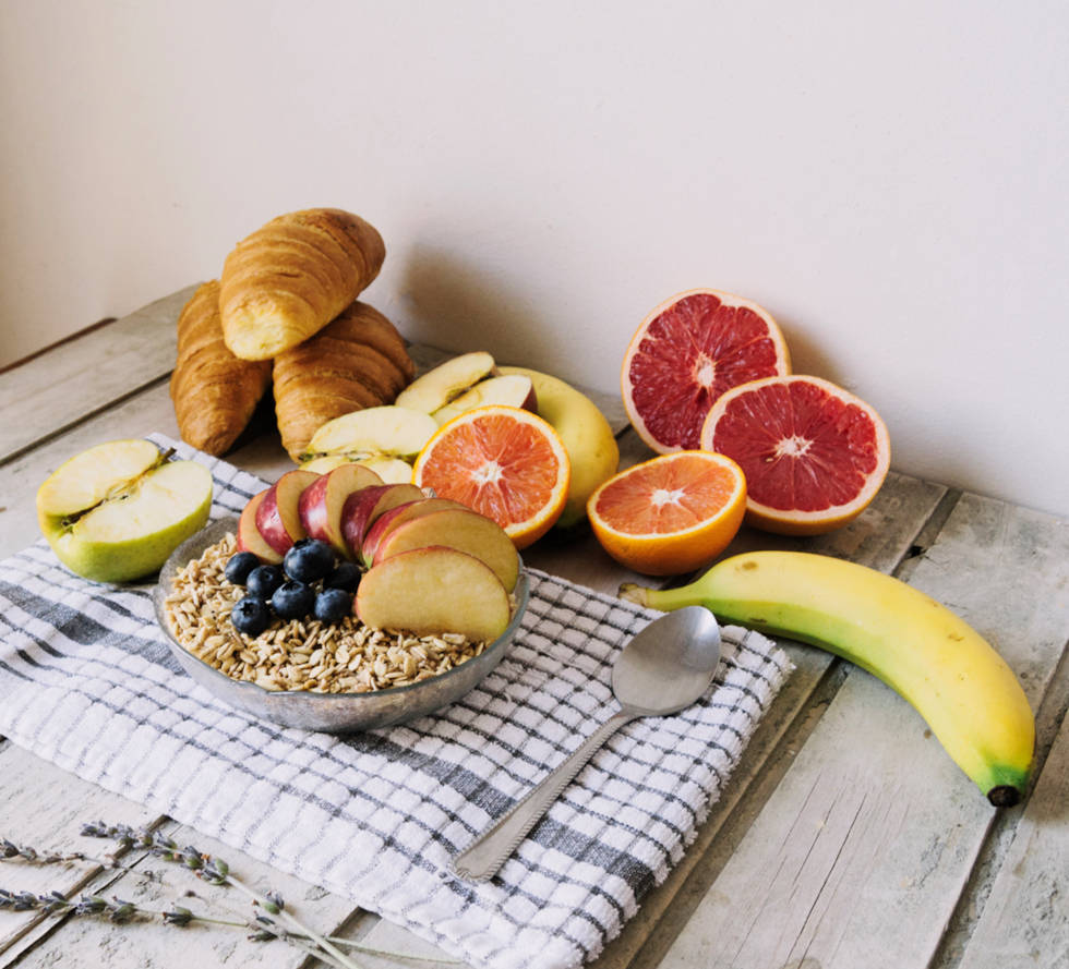Healthy breakfast with a bowl of oats topped with fruit, surrounded by fresh grapefruit, apple slices, bananas, and croissants on a wooden table.