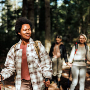 A group of hikers walking through a forest trail on a sunny day, with a woman in a plaid jacket leading the way and smiling.
