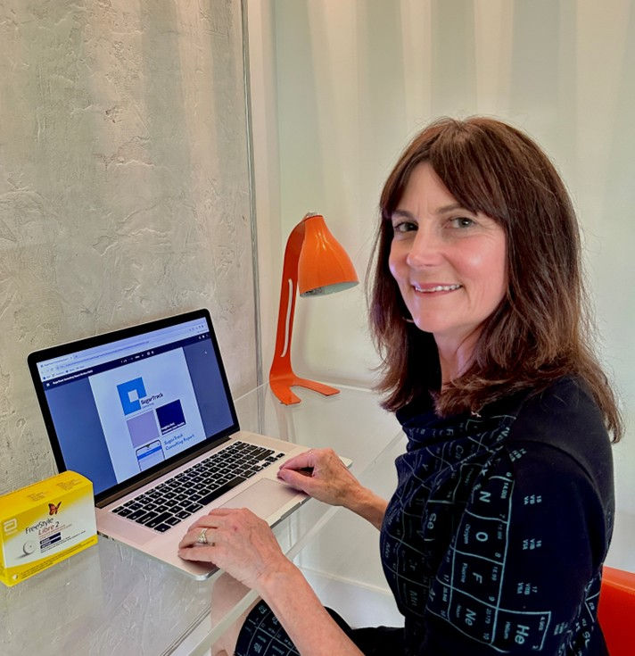 A woman sitting at a desk and smiling at the camera while working on a laptop. An orange desk lamp and a small yellow product box sit beside her.